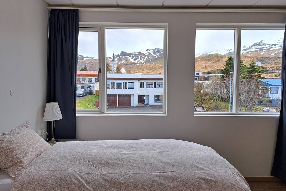 Un dormitorio con una cama grande, dos ventanas con vistas a un pueblo y montañas, una lámpara y una cortina.