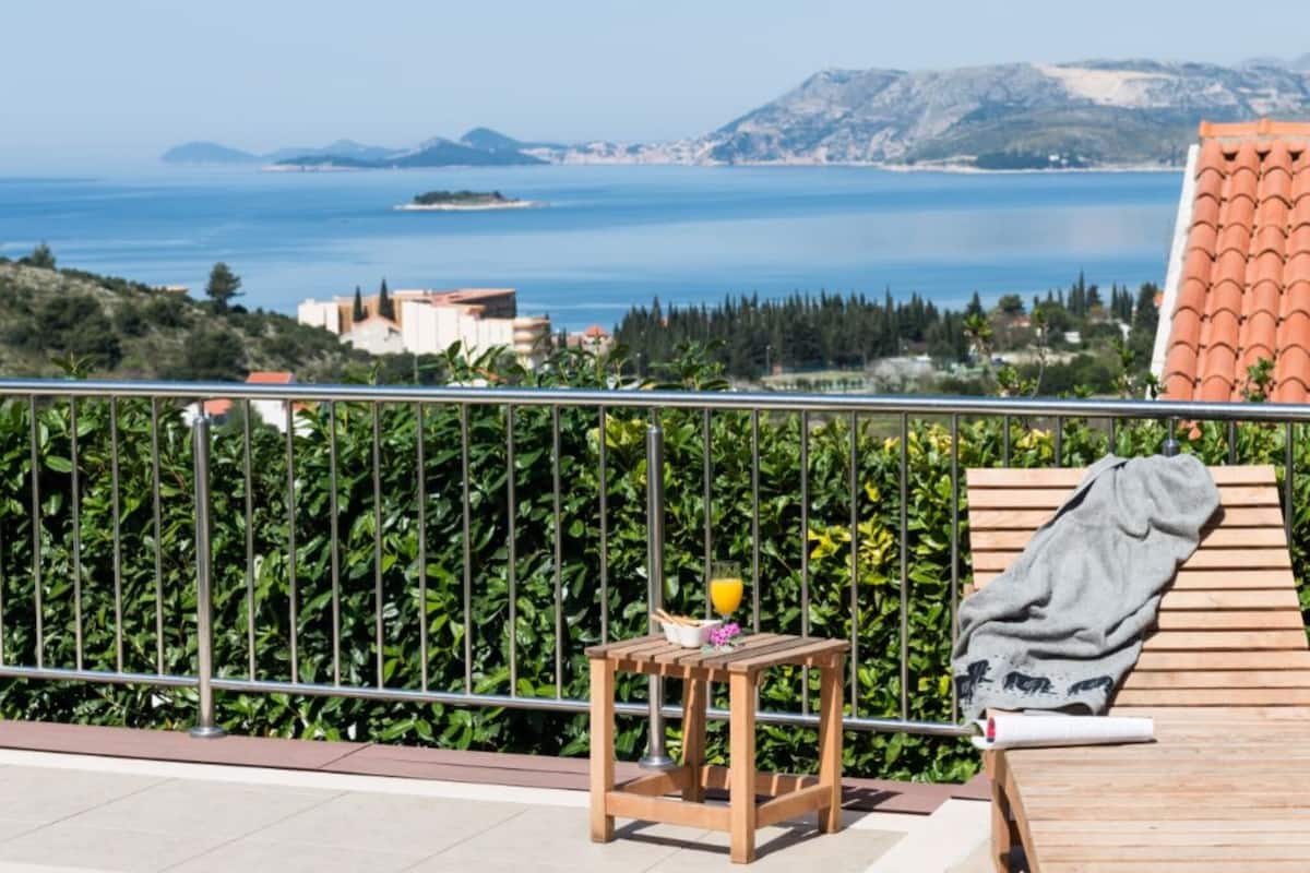 A balcony with a view of the sea, a wooden table with a glass of orange juice, and a towel on a chair.