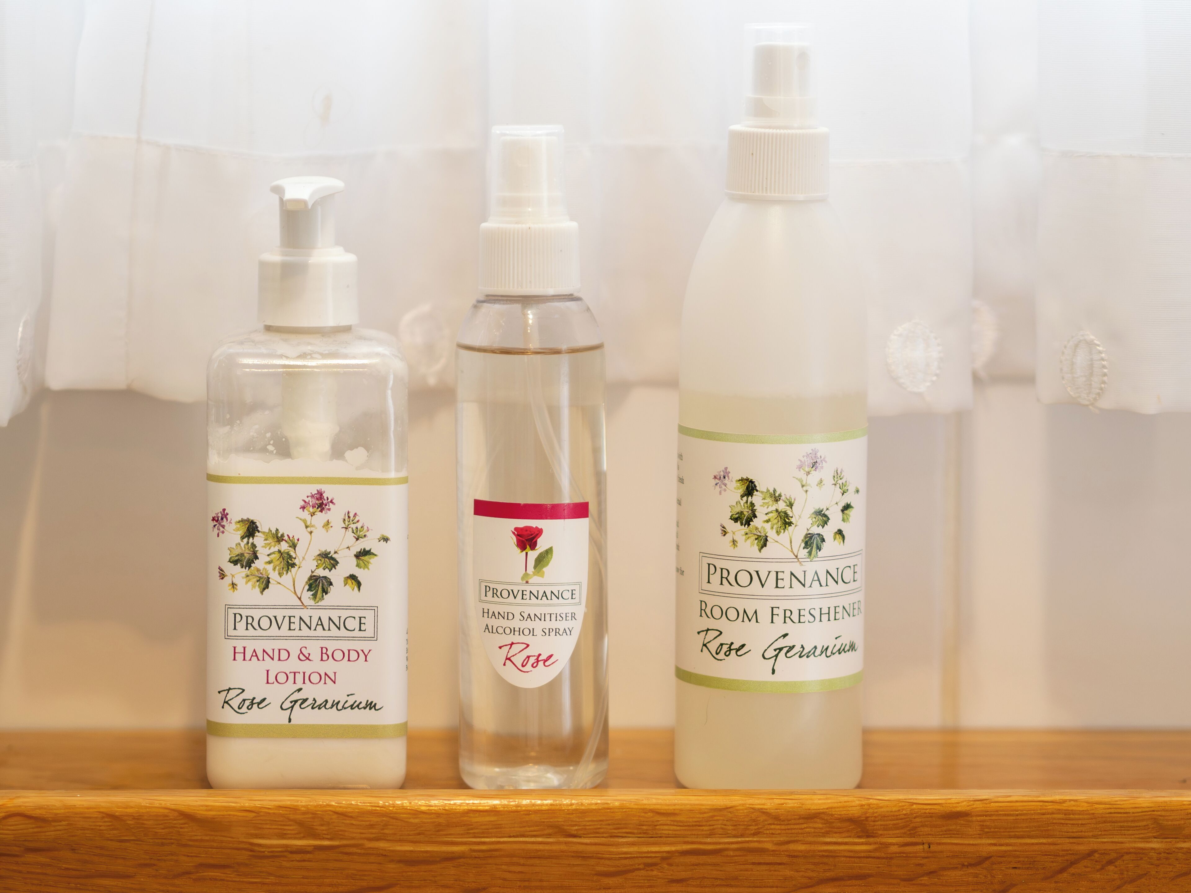 Three bottles of Provenance hand and body lotion, hand sanitizer, and room freshener on a wooden shelf.
