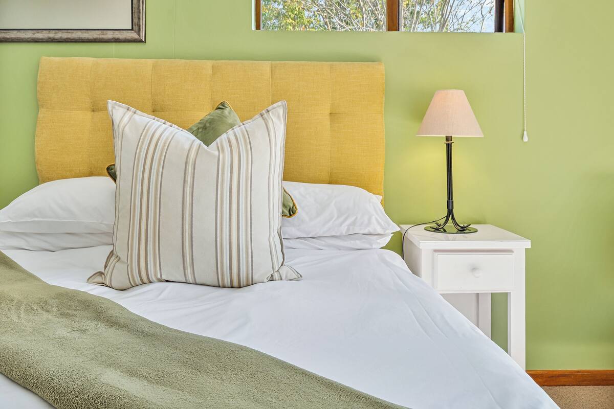 Uma cama bem arrumada com almofadas listradas, um cobertor verde e uma luminária de cabeceira sobre um criado-mudo branco.