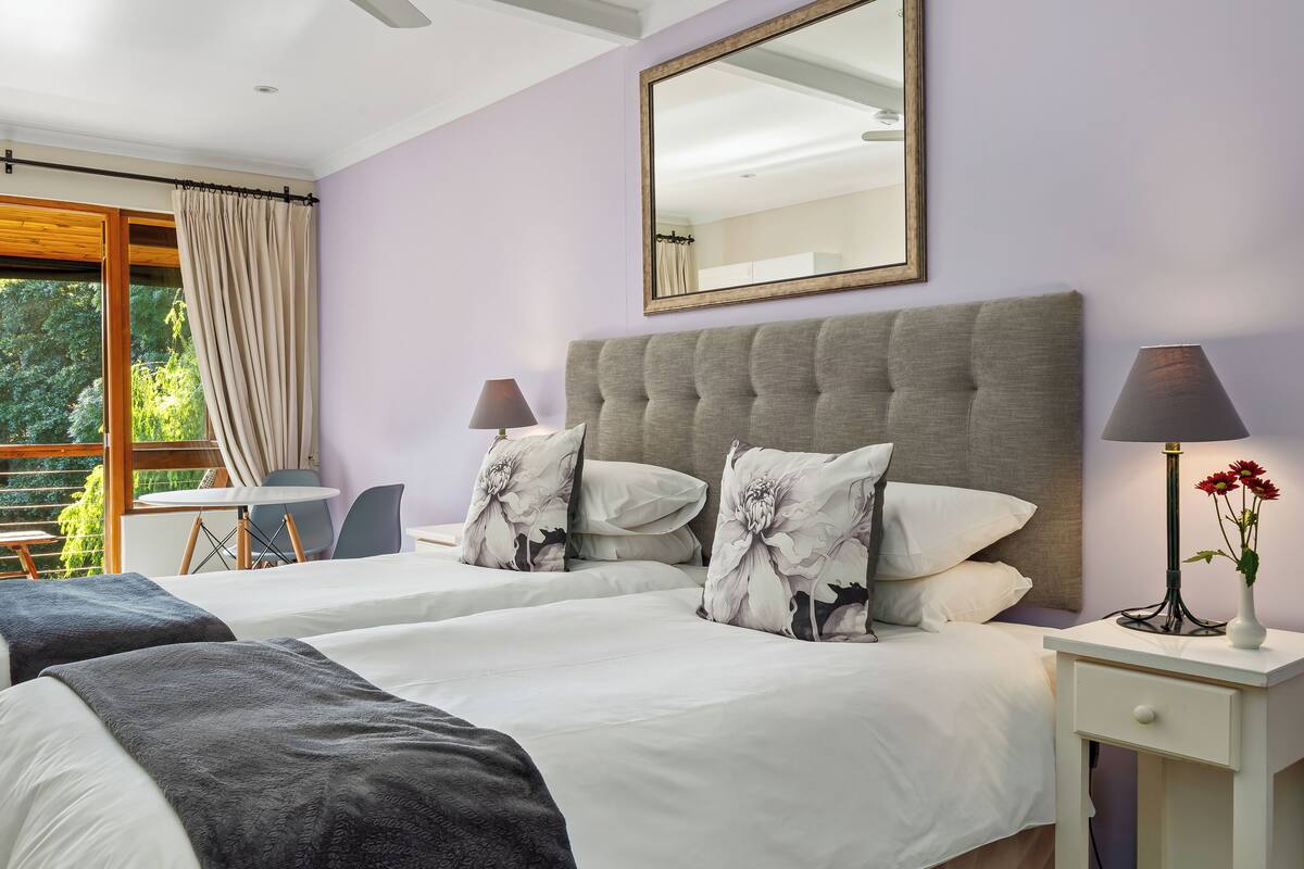 Quarto de hotel com duas camas, uma janela grande com cortinas, mesa de cabeceira com abajur e um espelho na parede.