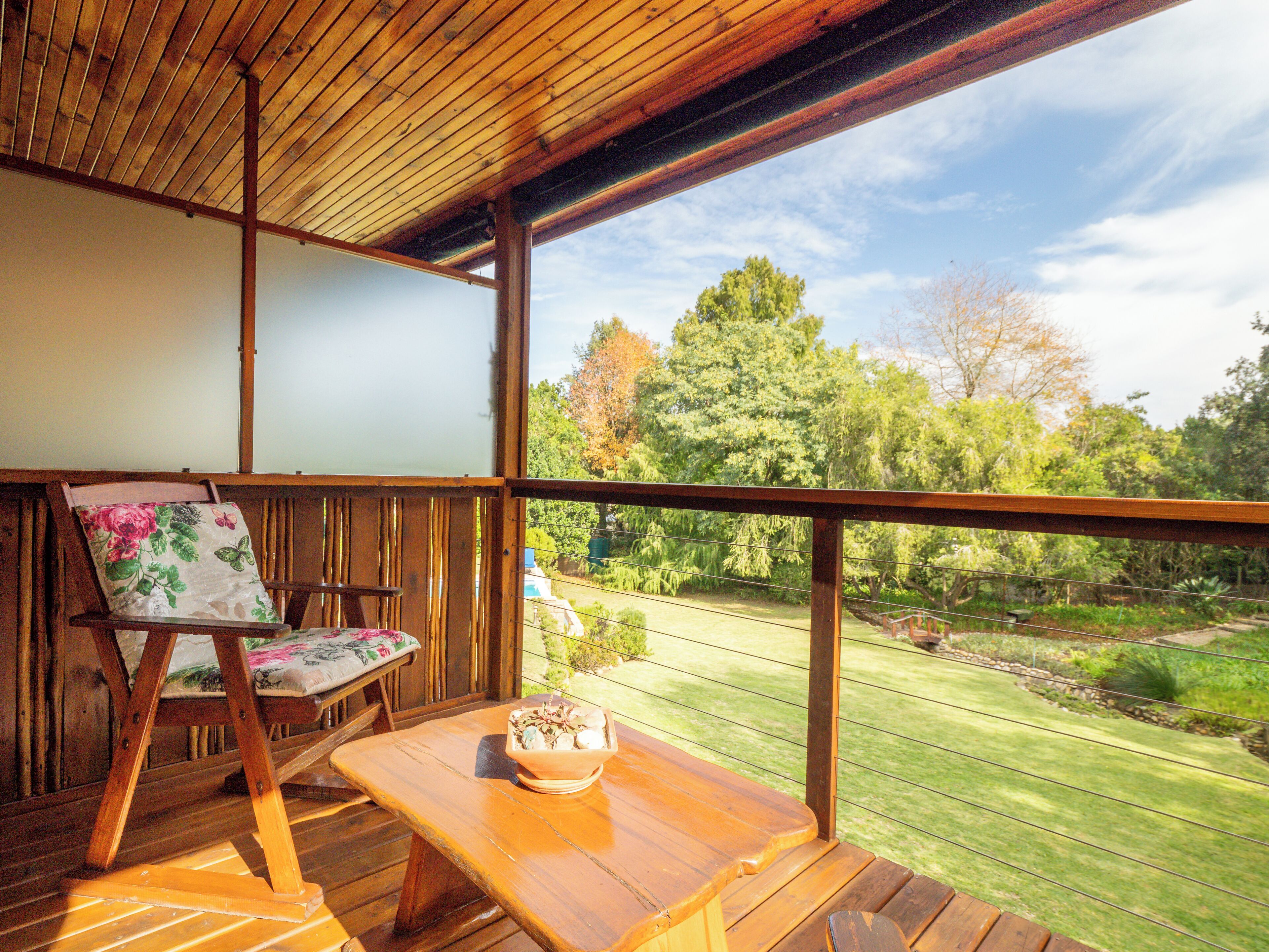 A wooden deck with a table and chair, a view of a garden with trees and a lawn.