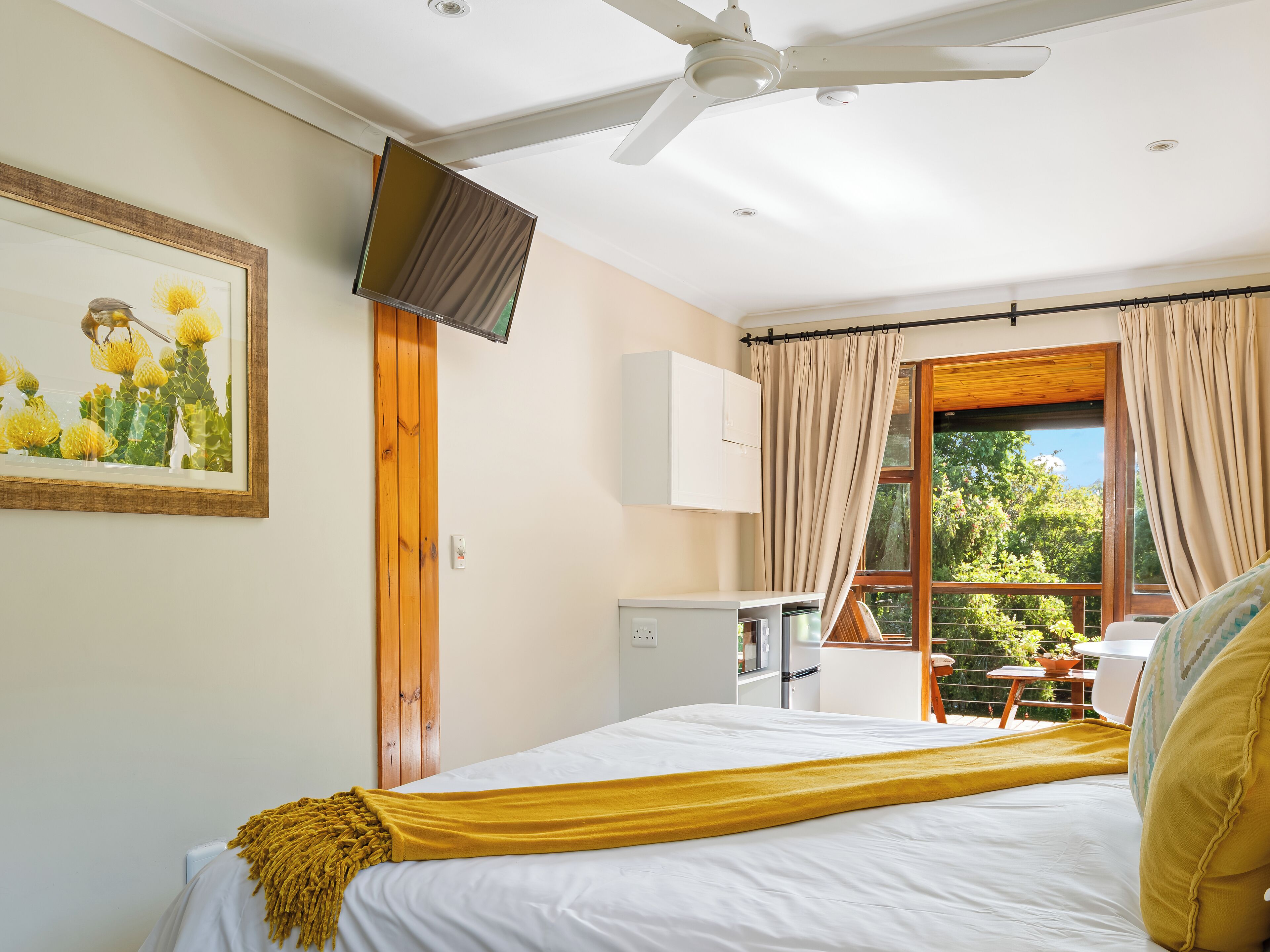 A bedroom with a bed, a ceiling fan, a TV mounted on the wall, a window with curtains, and a view of greenery outside.