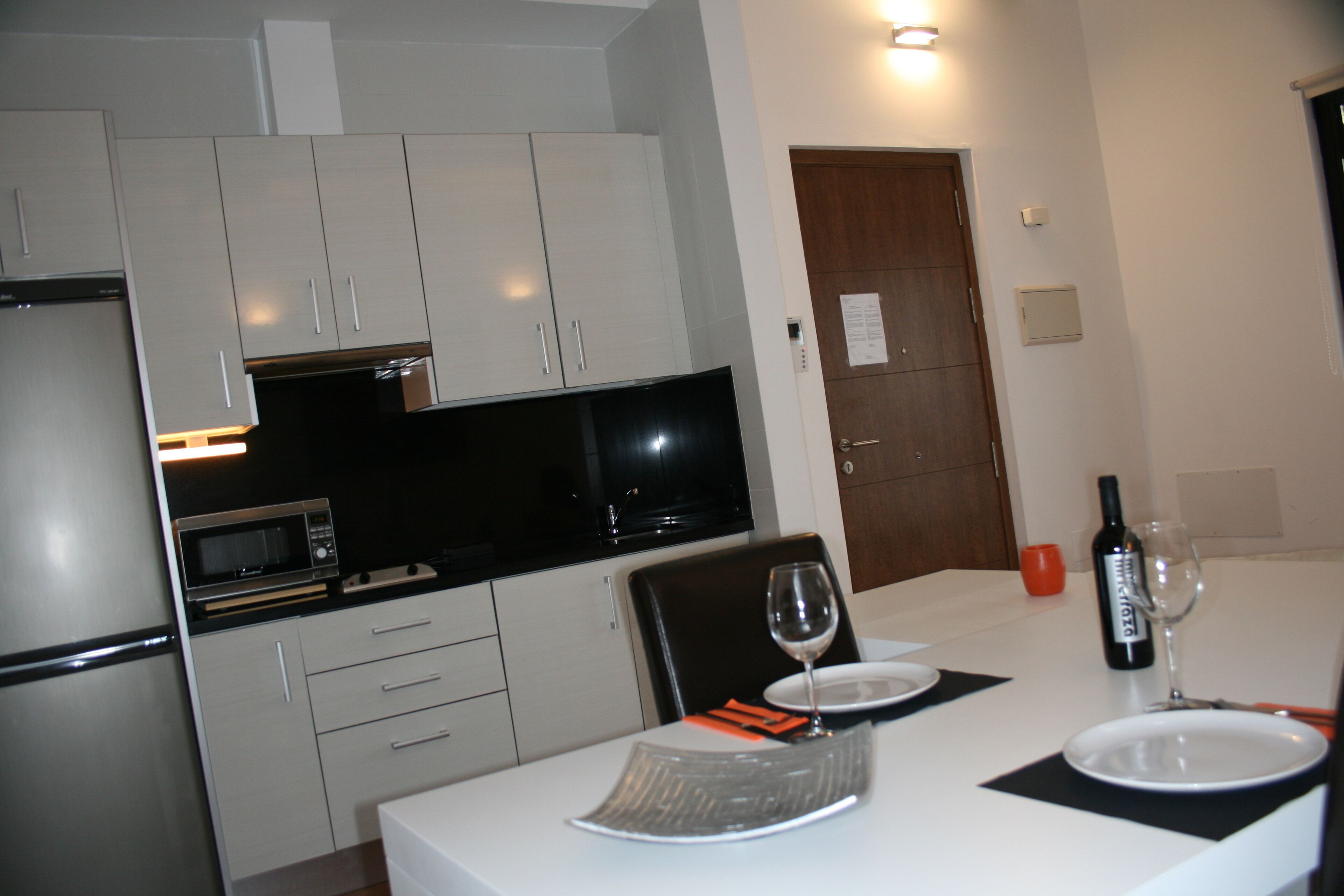 Een moderne keuken met witte kasten, een magnetron en een zwart aanrecht. Er staat een eettafel met een wijnglas en een fles wijn, en een deur op de achtergrond.