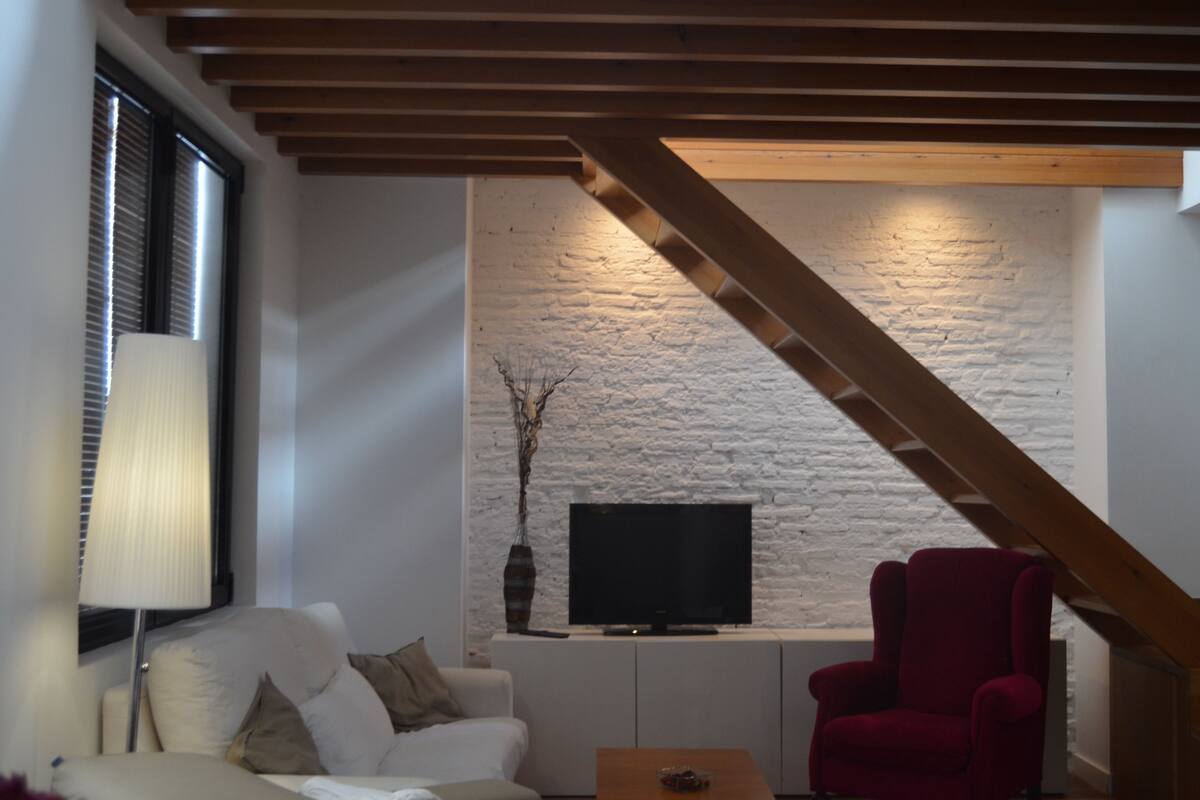 A modern living room with a white brick wall, a flat-screen TV, a red armchair, and a wooden staircase.