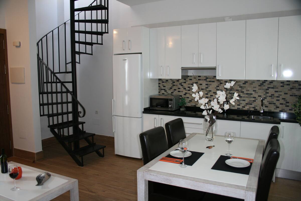 A modern kitchen with white cabinets, a black staircase, and a dining area with a white table set for two.
