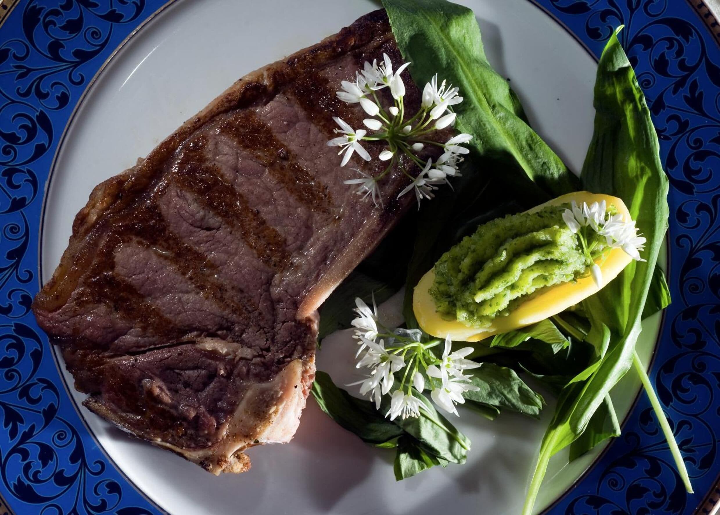 A plate with a steak, a slice of avocado, and some greens with flowers.