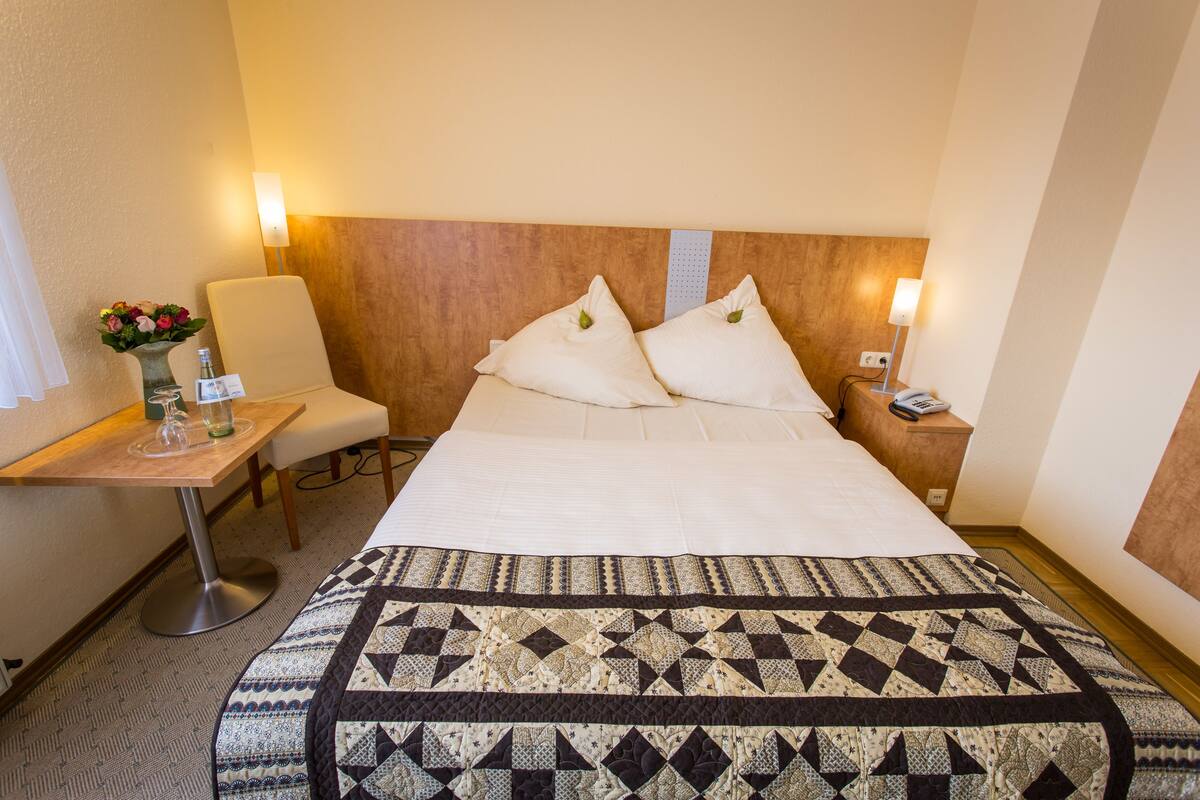 Ein Hotelzimmer mit einem großen Bett, einem Nachttisch mit Lampe, einem Sessel und einem kleinen Tisch mit Blumenvase.