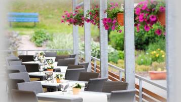Un café en plein air avec des meubles en osier, des tables recouvertes de nappes blanches et des paniers de fleurs suspendus.