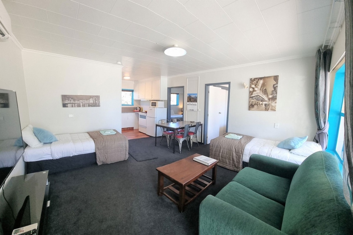A compact living space with a sofa, a green armchair, a small table, and a kitchenette in the background.