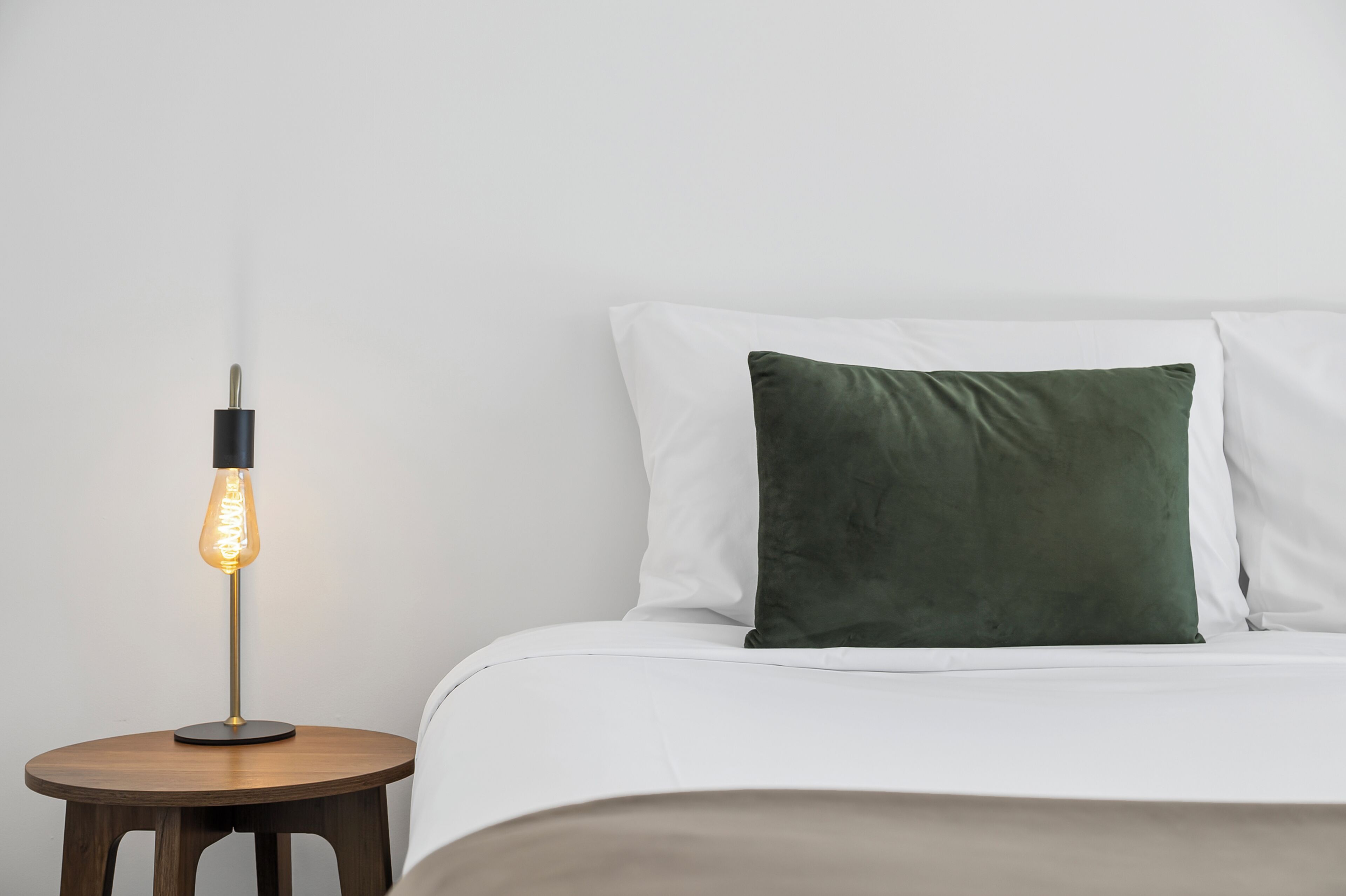 Una cama bien hecha con sábanas blancas y un almohadón verde, una mesita de noche redonda de madera con una lámpara moderna, y una pared blanca lisa.