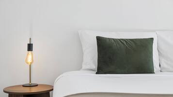 Una cama bien hecha con sábanas blancas y un almohadón verde, una mesita de noche redonda de madera con una lámpara moderna, y una pared blanca lisa.