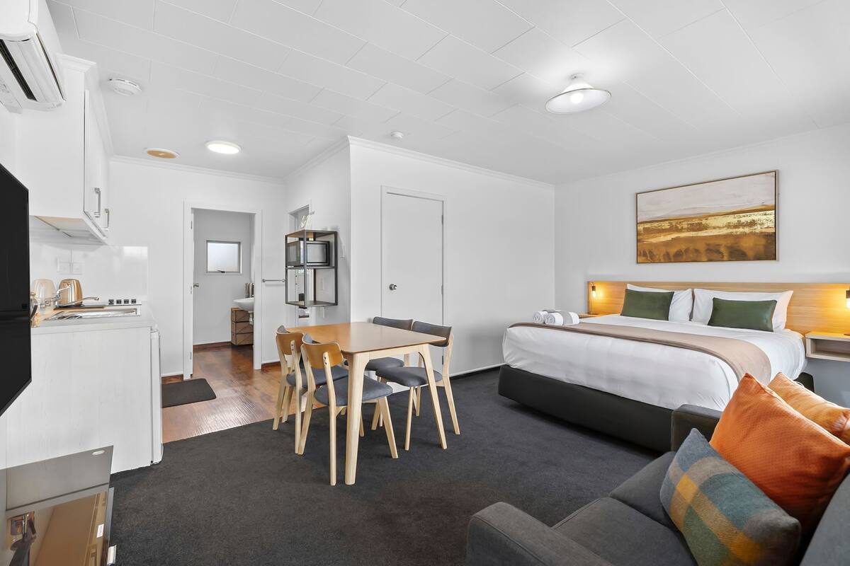 Une chambre d’hôtel moderne comprenant un lit, un canapé, une table à manger avec des chaises, un coin cuisine et un tableau au mur.