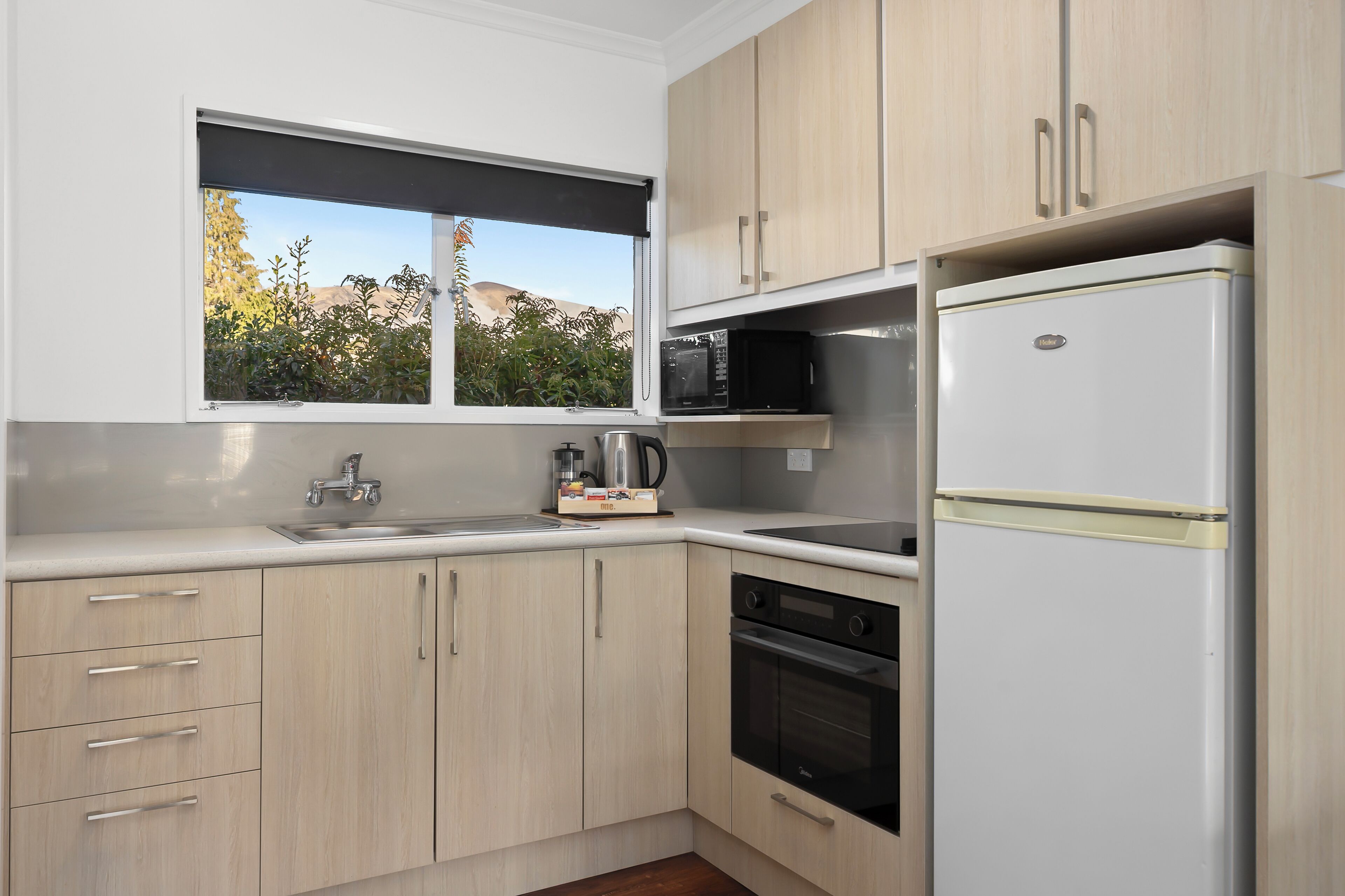 A modern kitchen with light wood cabinets, a white refrigerator, a microwave, and a window with a view of trees and mountains.