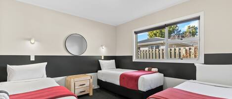 Habitación de hotel con tres camas, una mesita de noche de madera, un espejo redondo y una ventana que da a una valla y árboles.