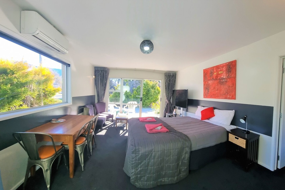 A hotel room with a bed, a desk, a chair, a TV, and a view of greenery outside.