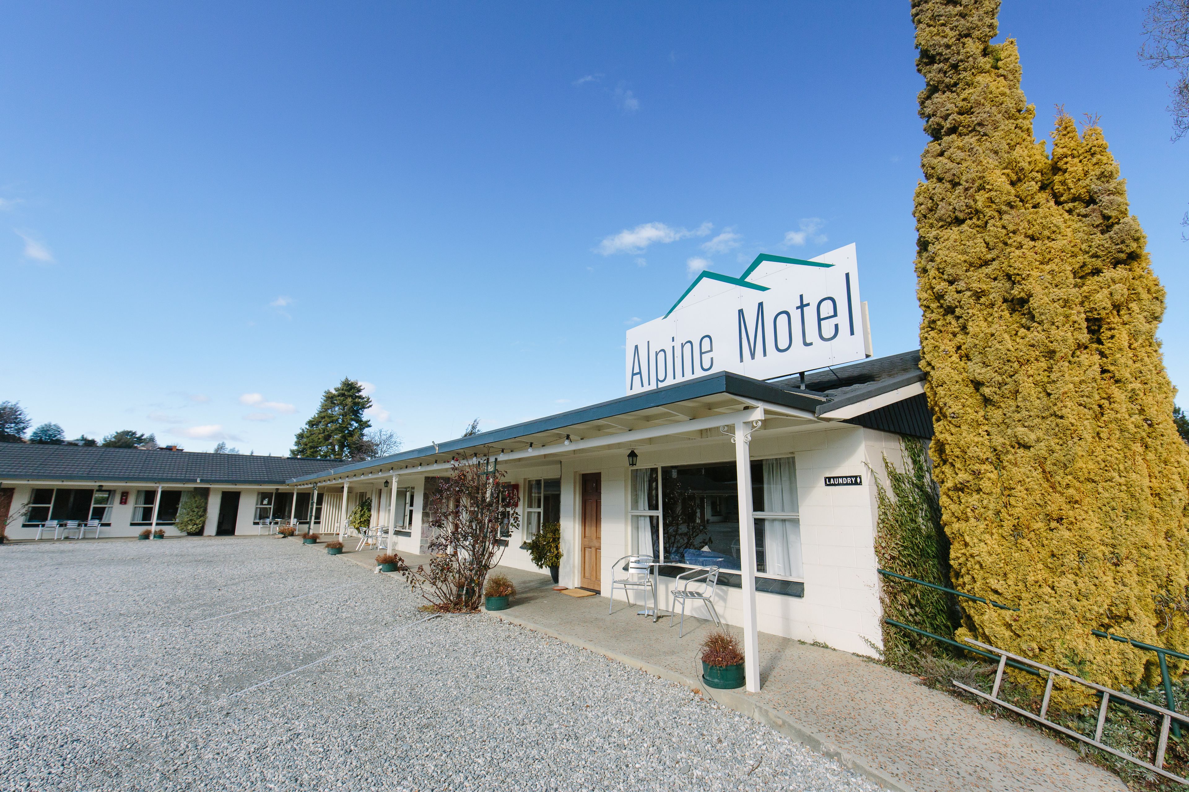 A motel with a sign reading "Alpine Motel," a covered front porch, and a gravel parking area.
