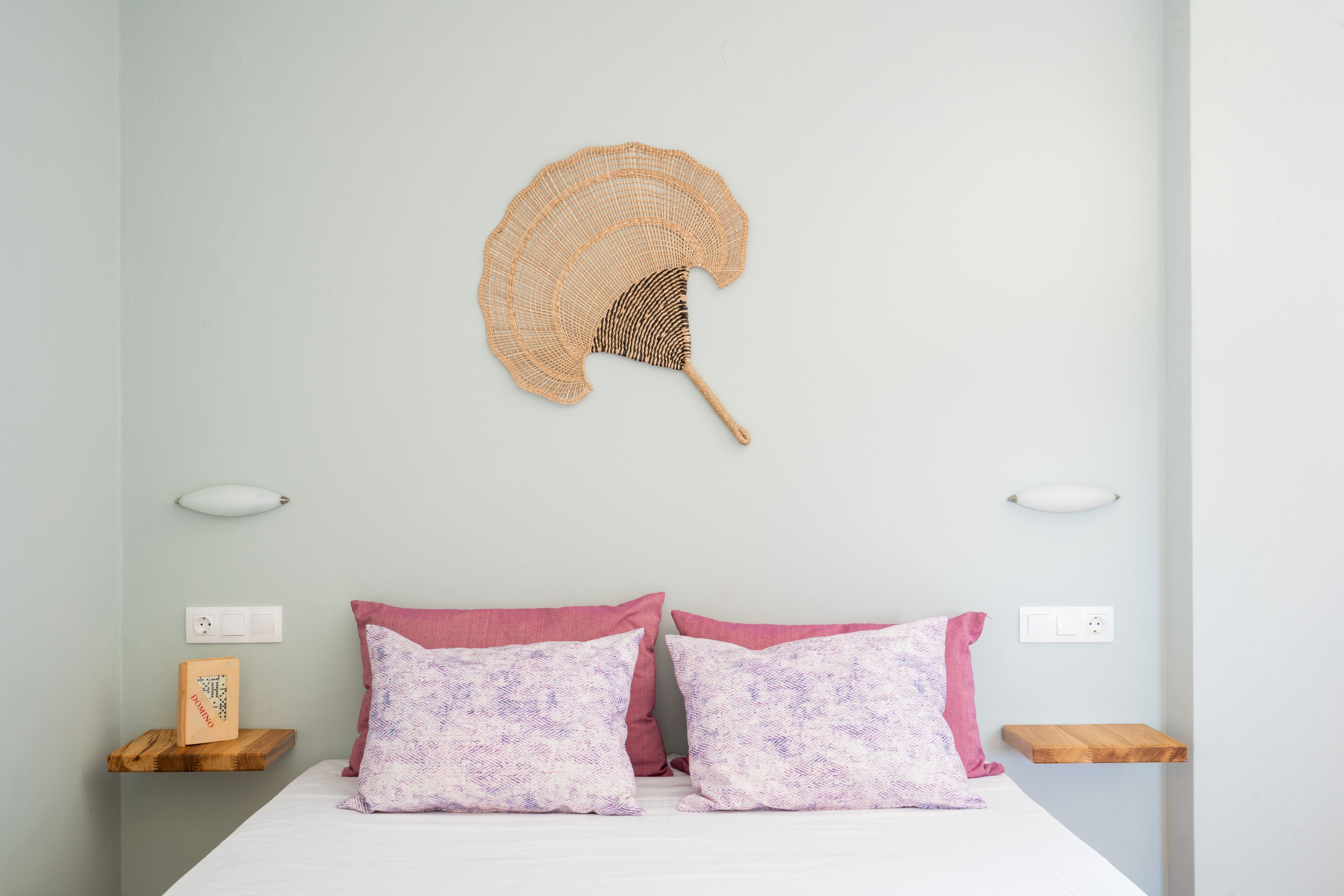 Een bed met twee kussens, een nachtkastje en een ventilator aan de muur.
