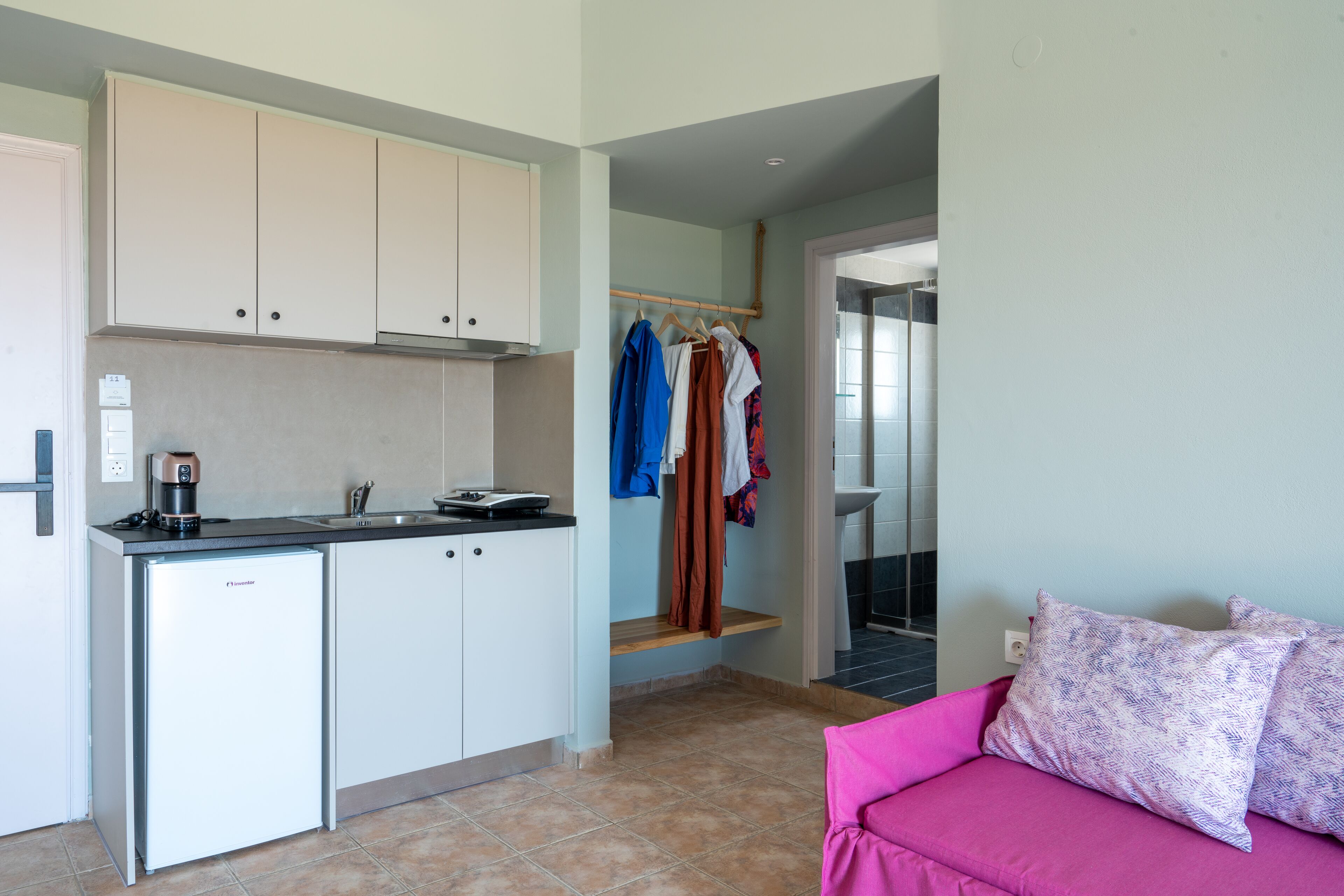 A compact kitchen with a refrigerator, a coffee maker, and a sink. There is a small seating area with a pink couch and patterned pillows.