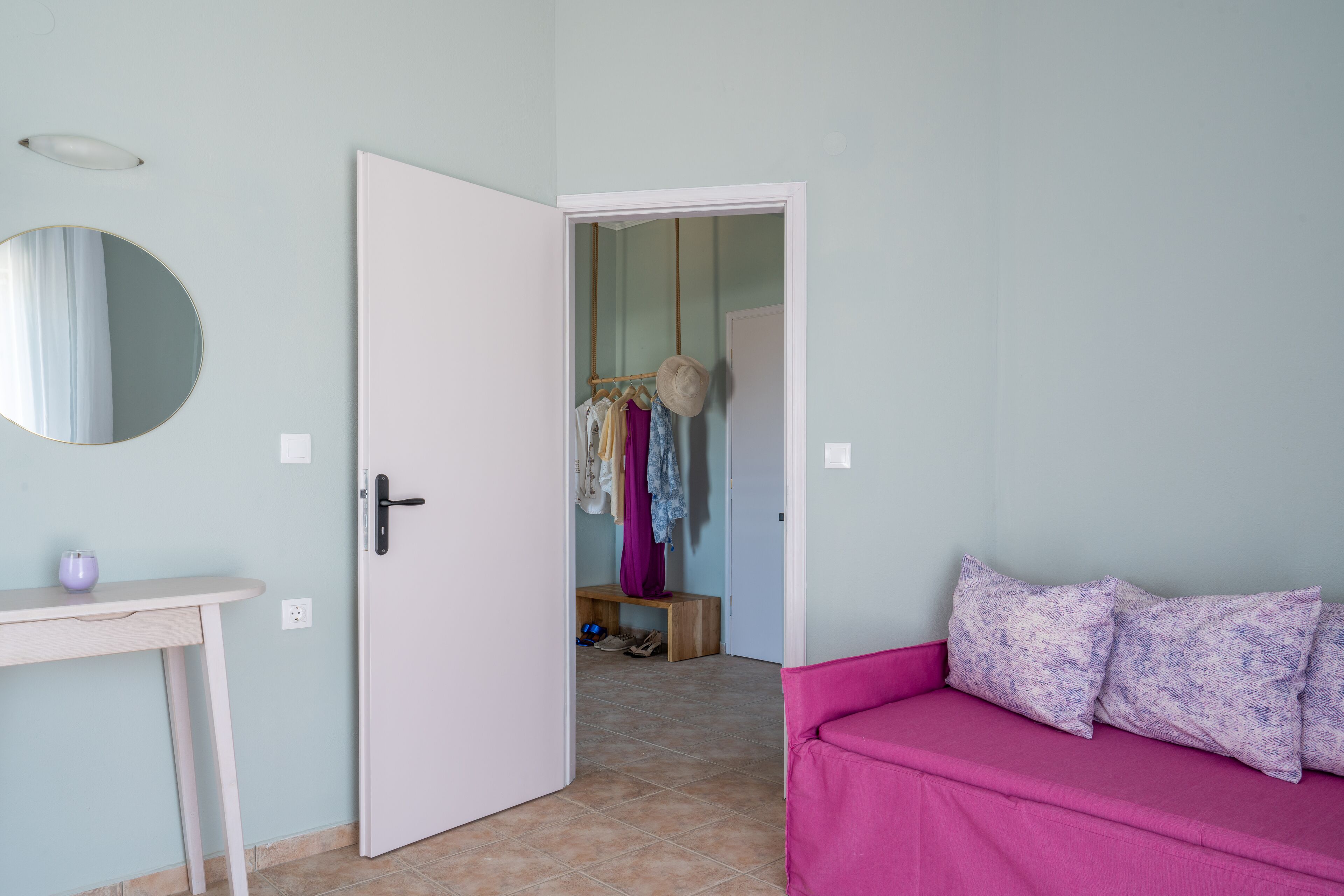 Quarto com sofá rosa, porta branca, espelho e um cabideiro com várias peças de roupa.
