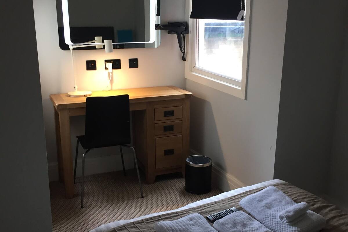 A bedroom with a bed, a desk with a chair, a mirror, a window, and a trash can.