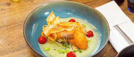 A blue bowl with a fish dish, garnished with cherry tomatoes and green peas, on a wooden table.