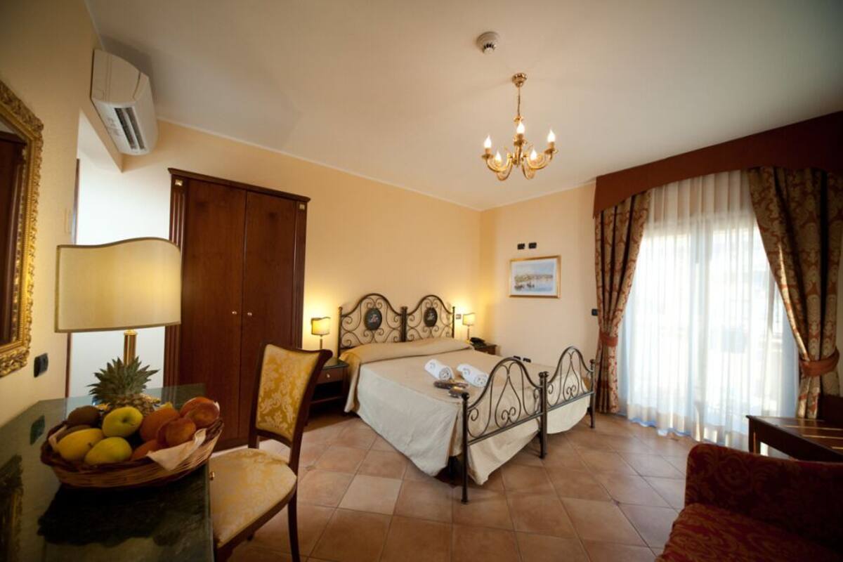A hotel room with a bed, a chair, a table with fruit, a lamp, and a chandelier.