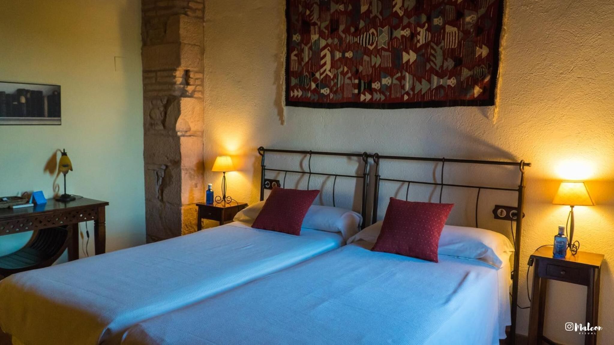 A double bed with two pillows, a nightstand with a lamp, a stone wall, and a tapestry on the wall.