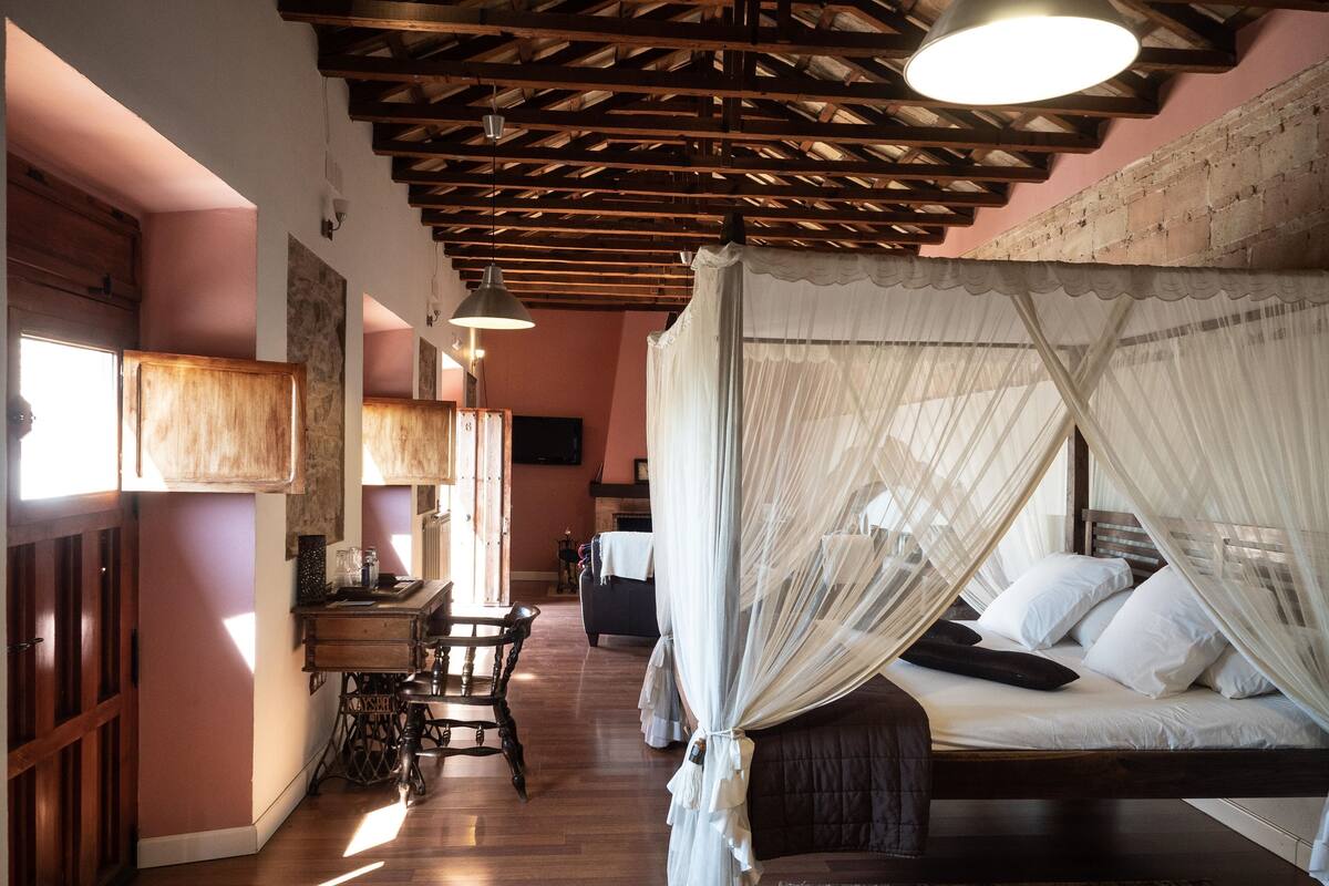 A bedroom with a canopy bed, wooden beams on the ceiling, a desk with a chair, and a television on the wall.
