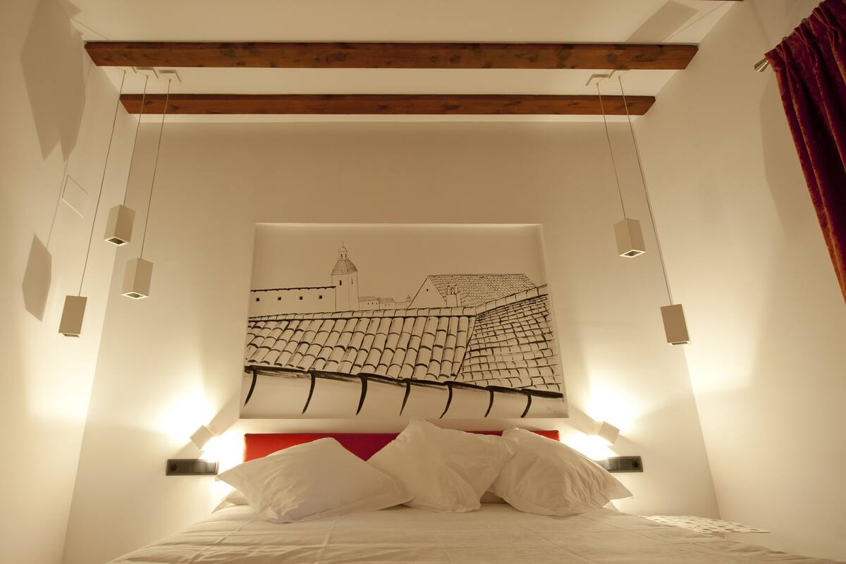 A bedroom with a bed, white bedding, a red headboard, and a wall-mounted artwork of a tiled roof.