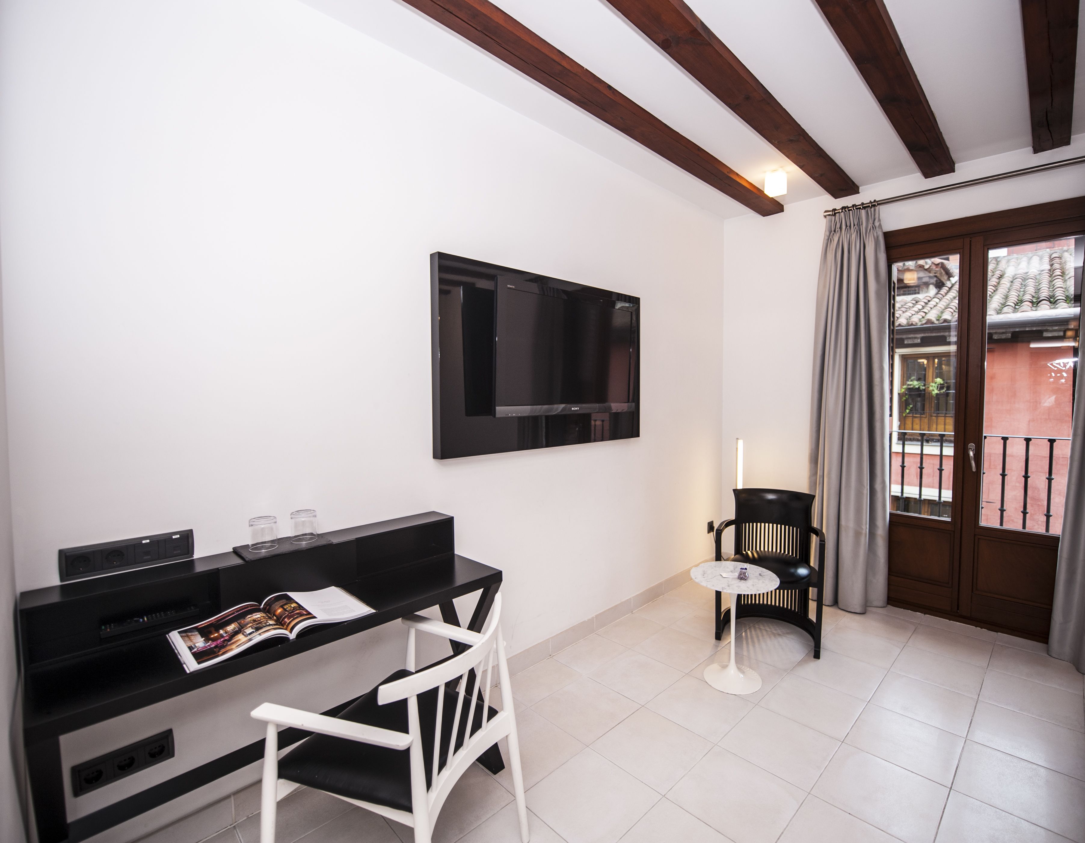 A modern room with a black TV on the wall, a white chair, a small round table, and a balcony with a view of buildings.