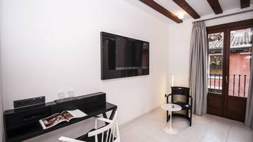 A modern room with a black TV on the wall, a white chair, a small round table, and a balcony with a view of buildings.