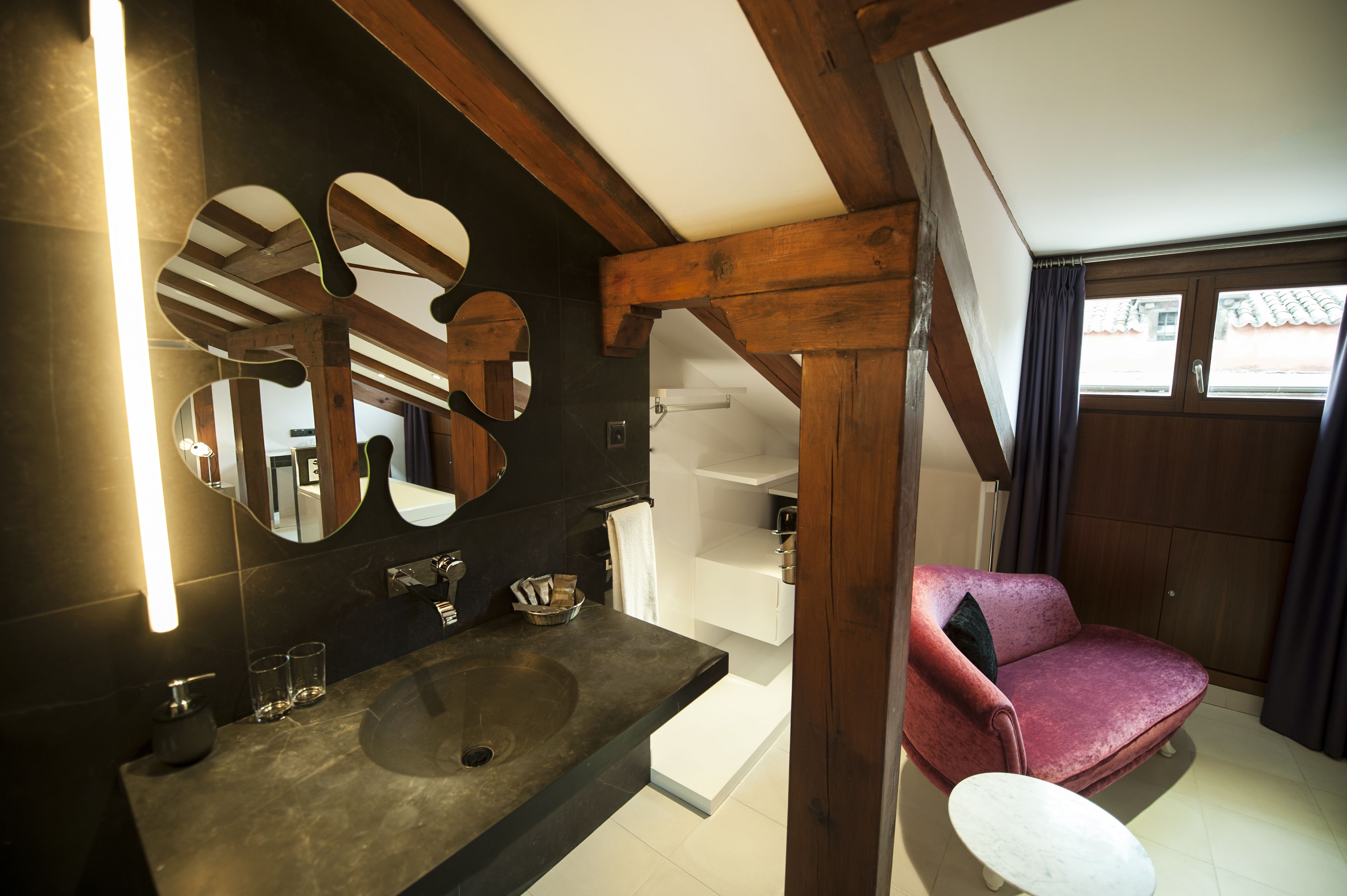 Un baño con un espejo grande, vigas de madera, un lavamanos y una silla roja.