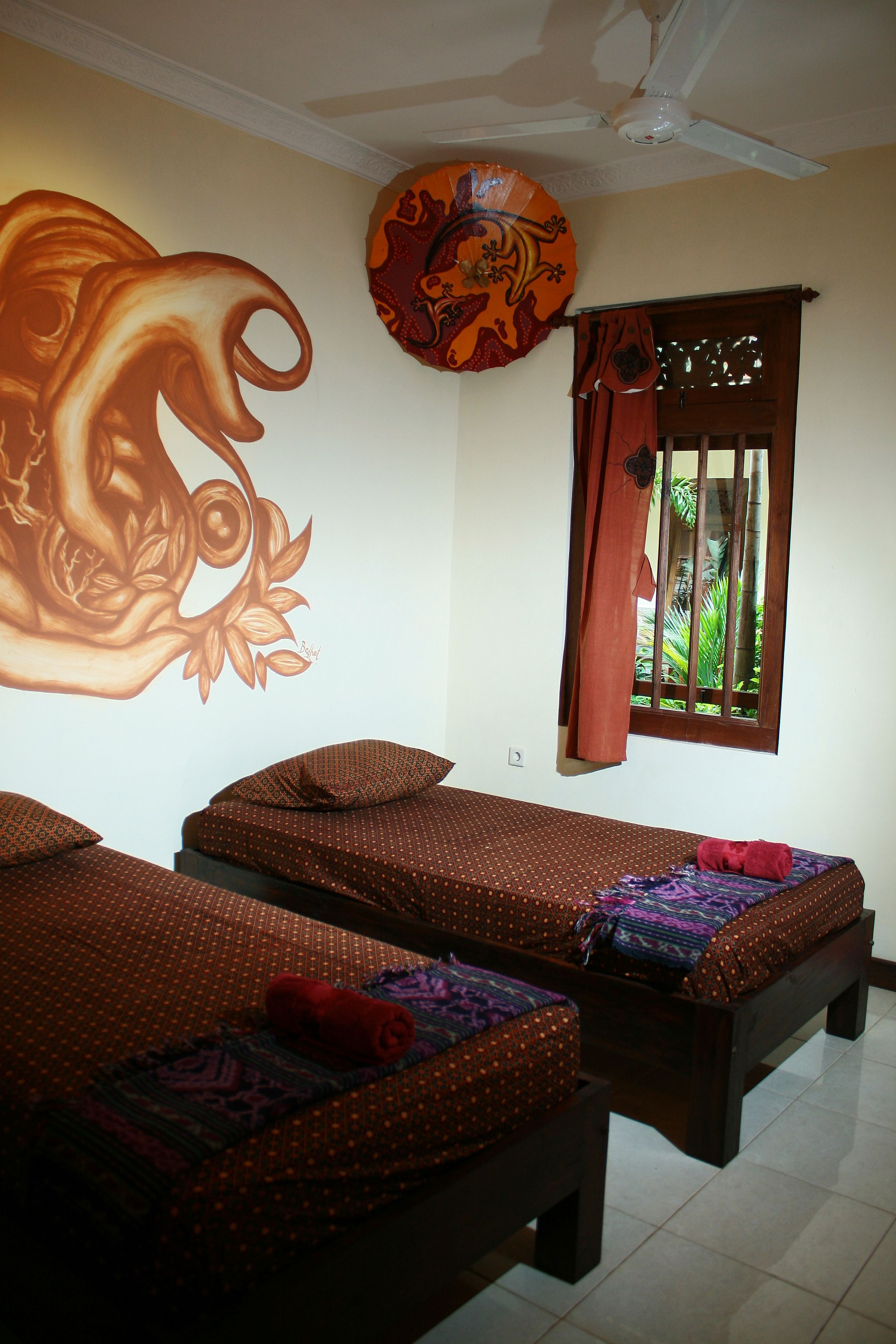 Una habitación con dos camas individuales, ventilador de techo, ventana con cortinas y un mural en la pared.