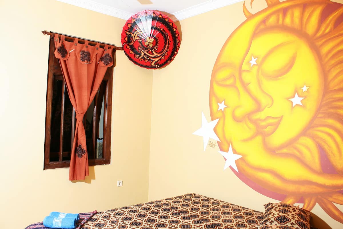 Une pièce avec un lit, une fenêtre avec des rideaux et un motif décoratif représentant un soleil sur un mur.