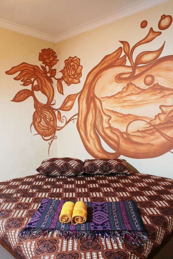 A bed with patterned bedding, a wall mural, and two rolled towels. - Hotel 1001 Malam (Yogyakarta)