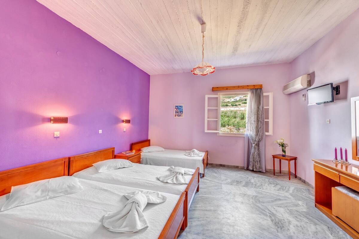 A hotel room with two beds, a wooden headboard, a window with curtains, a small table with a vase, and a TV on the wall.