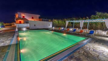 A swimming pool with lounge chairs and a covered area, surrounded by stone flooring and a white building with red accents.