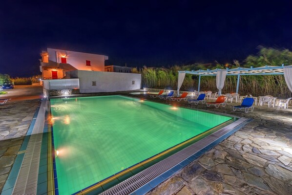 A swimming pool with lounge chairs and a covered area, surrounded by stone flooring and a white building with red accents.