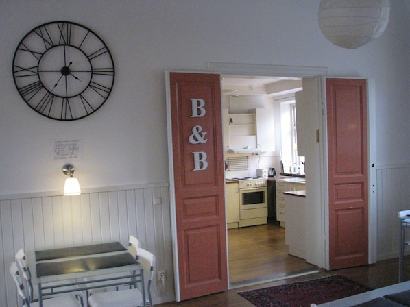Een kamer met een bed, een bureau en een wandklok. Een deur met 'B&B' erop leidt naar een keukenhoek.