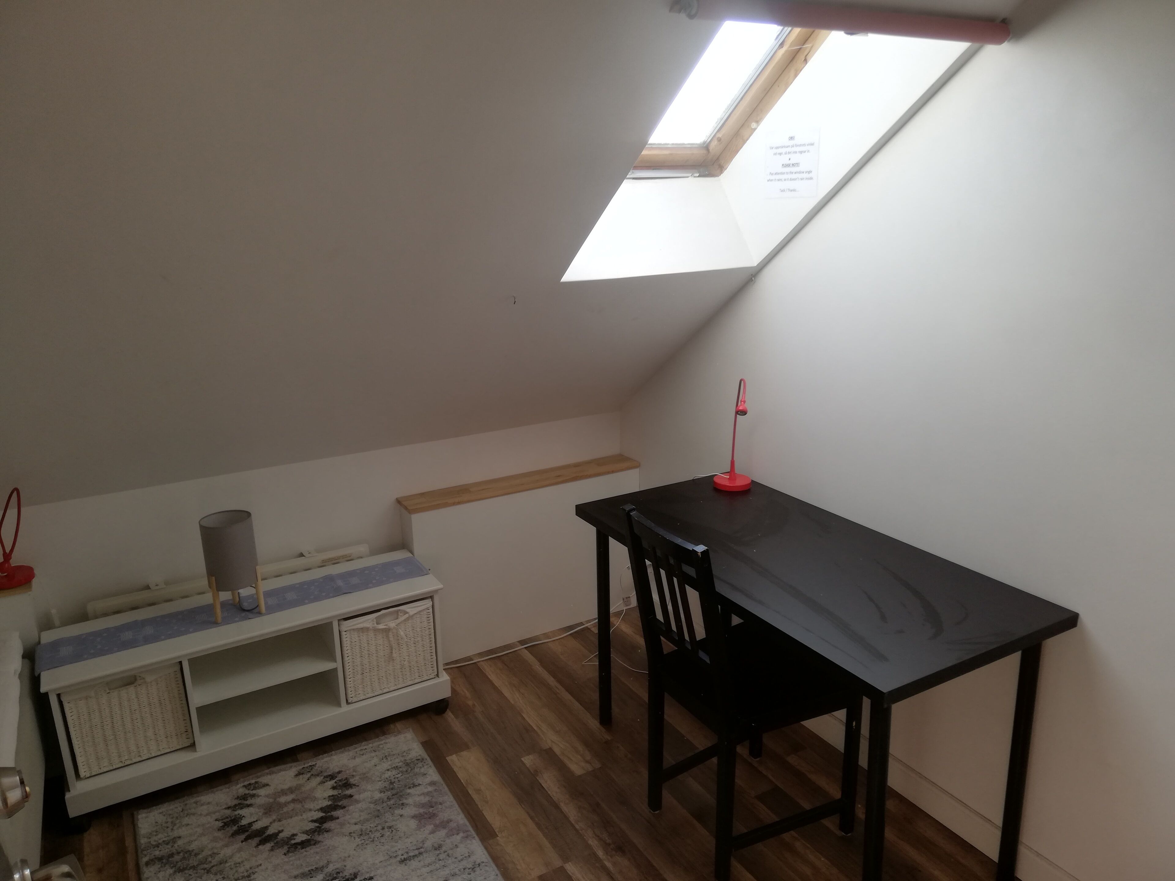 Een kleine kamer met schuine wanden, een dakraam, een bureau met stoel, een klein plankje en een opbergunit.