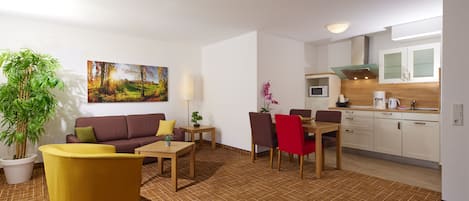 Una sala moderna con un sofá, sillas, una mesa de centro y una mesa de comedor. Se observa una zona de cocina con armarios y electrodomésticos. Una planta grande se ve en la esquina.