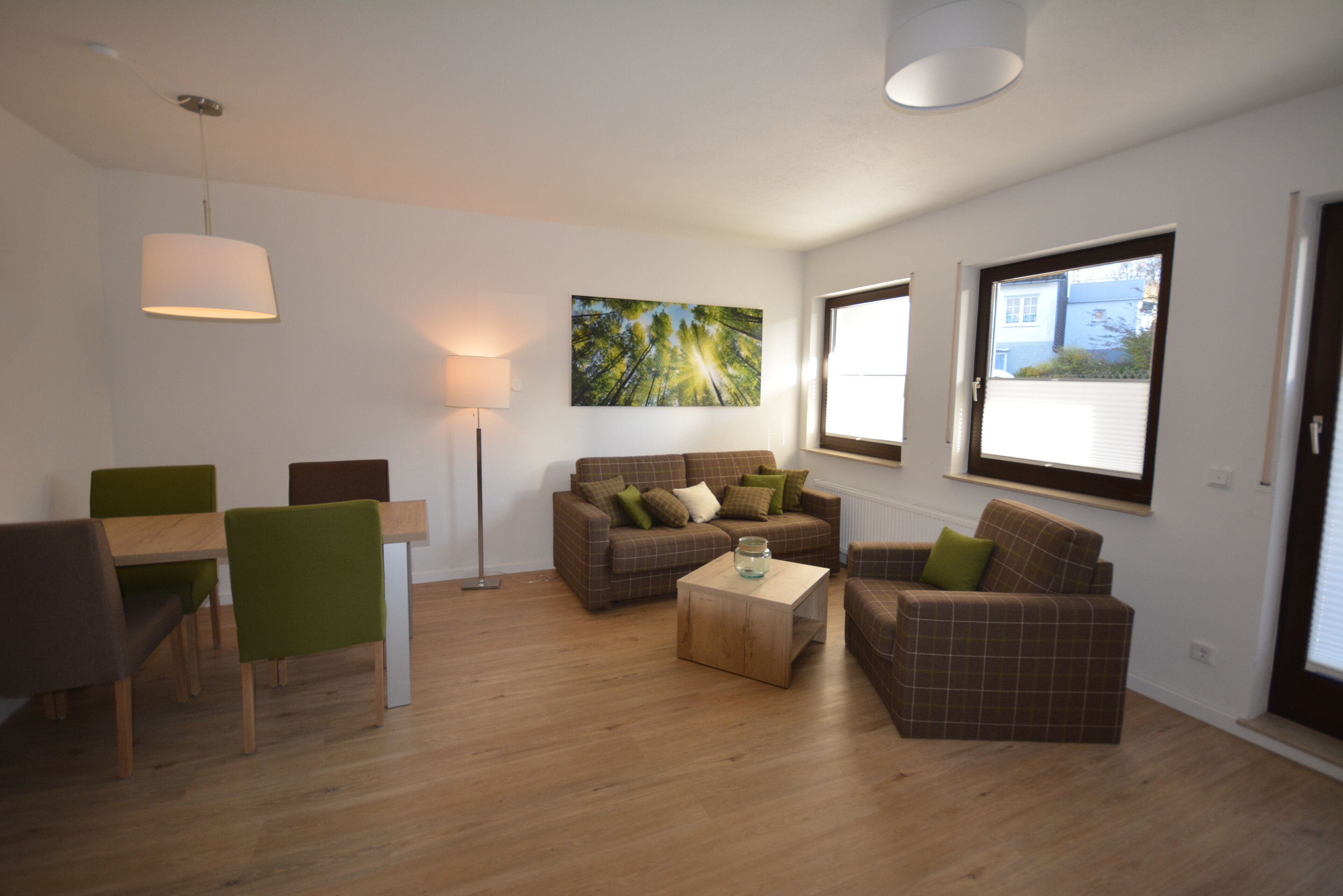 A modern living room with a sofa set, a dining table with chairs, a coffee table, and a floor lamp. There is a large window providing natural light and a view of the outside.