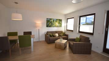 A modern living room with a sofa set, a dining table with chairs, a coffee table, and a floor lamp. There is a large window providing natural light and a view of the outside.