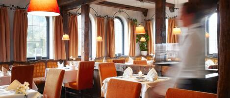A restaurant with tables set for dining, featuring a warm interior with wooden beams and large windows.