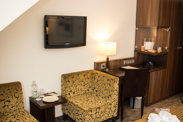 A hotel room with a sofa, a small table with a lamp, a television, a minibar, and a bathroom area. - AVITAL Resort Winterberg (Winterberg)