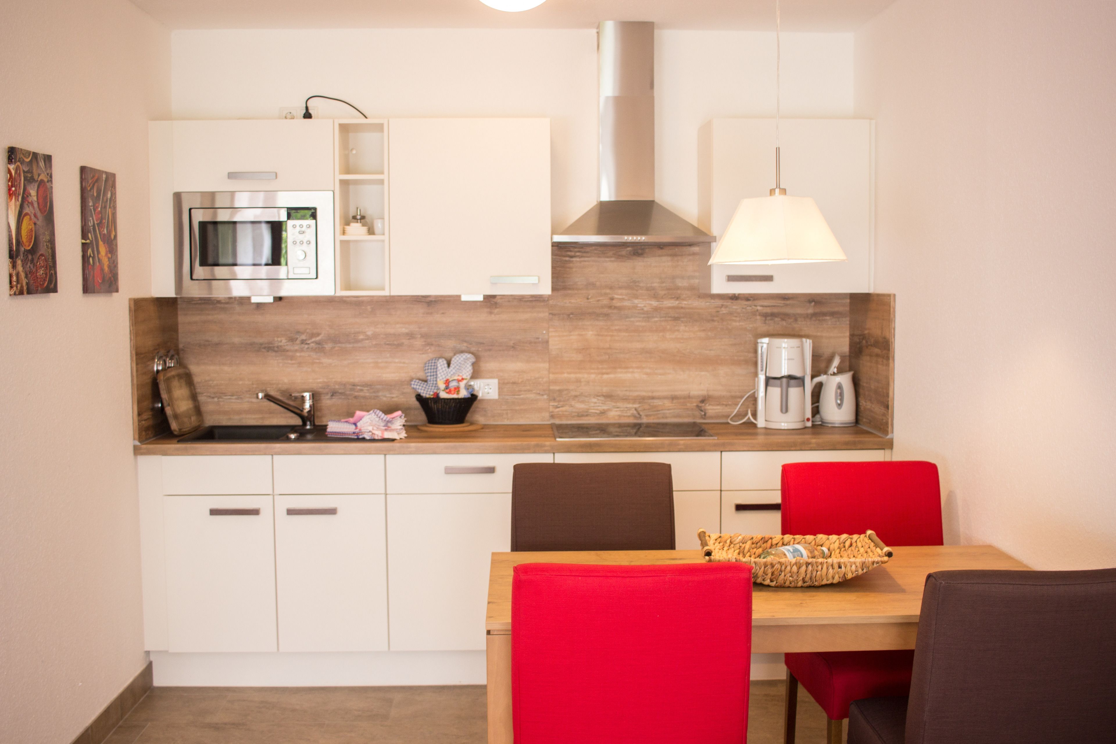 Una cocina moderna con armarios blancos, microondas, cafetera y un comedor con mesa de madera y sillas rojas.