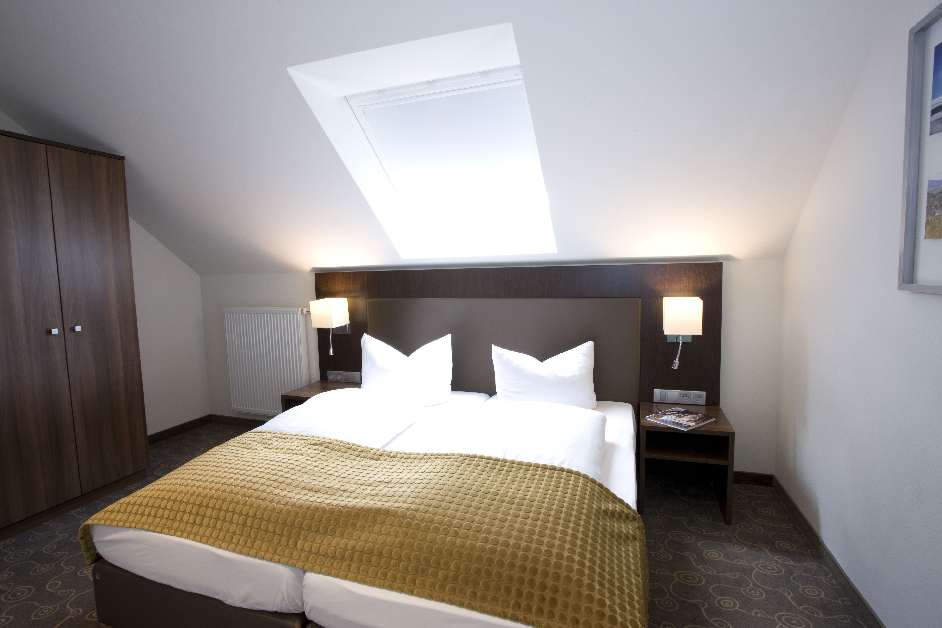 A hotel room with a bed, a skylight, a wardrobe, and a nightstand with a lamp.