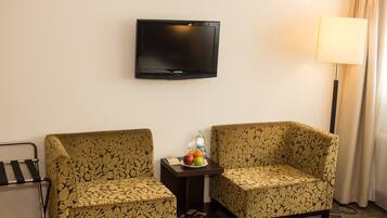 A hotel room with two patterned armchairs, a small table with fruit and a bottle, a flat-screen TV, a lamp, and a bed.