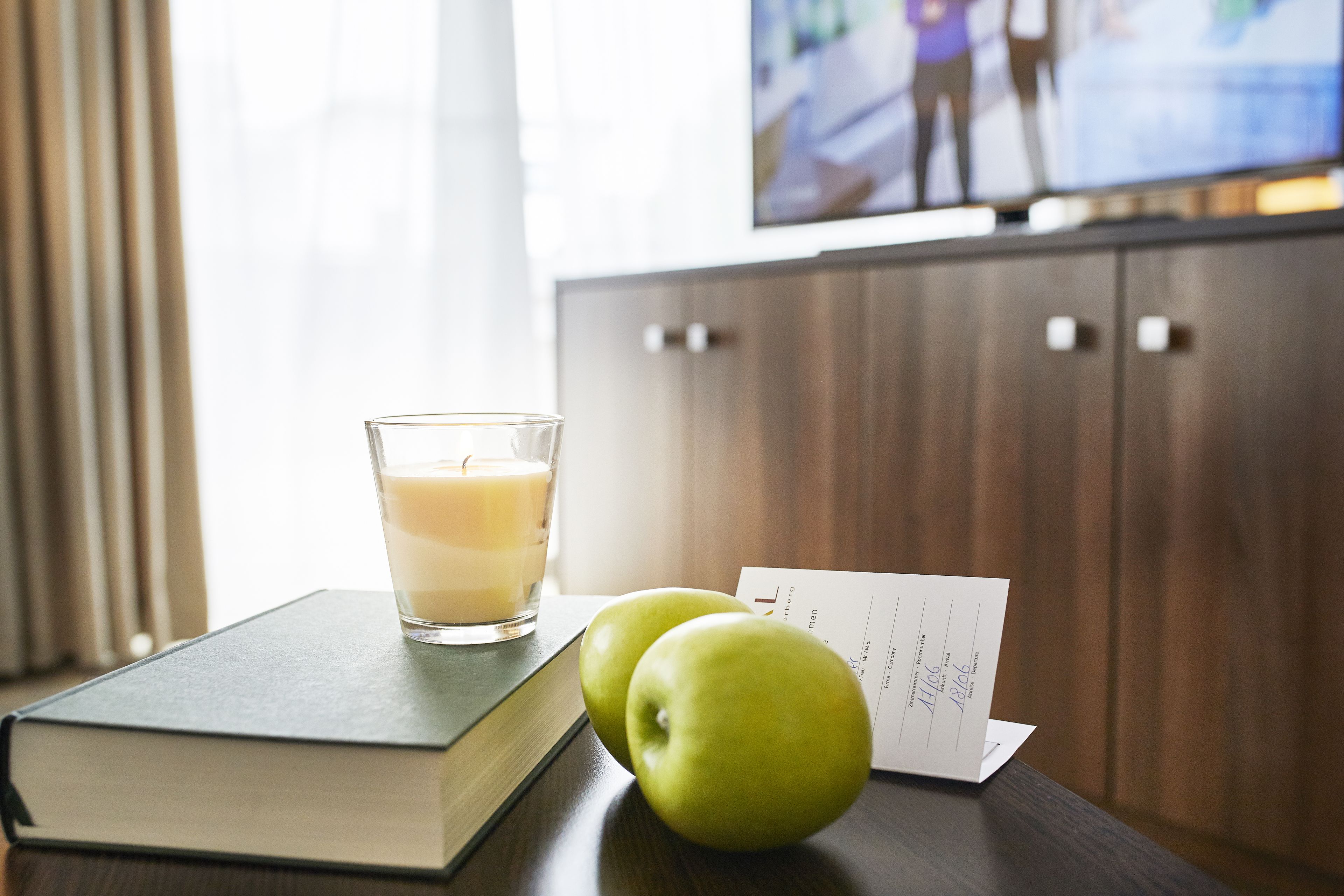 A book, a glass of milk, and two apples on a wooden table with a TV in the background.